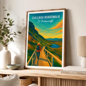 Puede incluir: Una lámina enmarcada con un paisaje estilizado del Cuilcagh Boardwalk en el condado de Fermanagh. La ilustración muestra a una persona con mochila caminando por un sendero de madera hacia un lago azul, rodeado de colinas verdes y un cielo naranja.