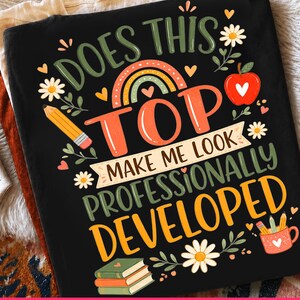 May include: A black t-shirt featuring a colorful graphic with the text DOES THIS TOP MAKE ME LOOK PROFESSIONALLY DEVELOPED. The design includes a rainbow, a red apple, a yellow pencil, a stack of books, a mug with pencils, and white flowers.