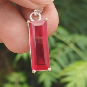 Puede incluir: Un colgante rectangular con una piedra preciosa de color rojo intenso facetada, engastada en un marco de metal plateado con cuatro garras en las esquinas y una anilla circular en la parte superior, con una superficie pulida.