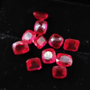 Puede incluir: Una colección de diez piedras preciosas rojas facetadas con un corte tipo cojín o rectangular. Las piedras exhiben un color rojo vibrante y translúcido con facetas internas visibles que reflejan la luz, dispuestas en un grupo sobre una superficie oscura.