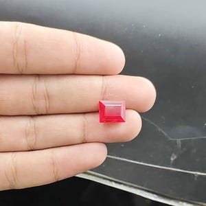 Può includere: Una gemma rossa dal taglio rettangolare con una superficie sfaccettata che riflette la luce. La pietra presenta bordi netti e definiti e una tonalità cremisi profonda e satura, mostrando una qualità trasparente e limpida nella struttura.