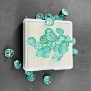 May include: A collection of round, faceted, translucent green gemstones with internal crackled textures. Many of the stones are clustered together on a square white foam display pad, while several individual stones are scattered around the base.