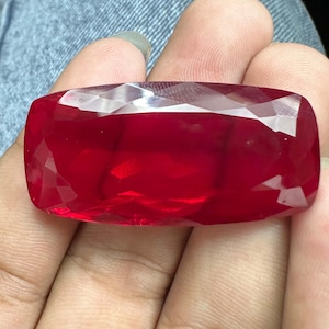 以下が含まれることがあります： 鮮やかで深い赤色をした、大きな長方形のファセットカット宝石。石の表面は研磨されており、複数の幾何学的なファセットが光を反射し、その細長い形状全体に輝きのある半透明の外観を作り出しています。