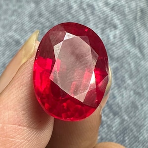Può includere: Una gemma dal taglio ovale con un colore rosso intenso e saturo, caratterizzata da una superficie sfaccettata che riflette la luce. La pietra mostra una qualità trasparente e limpida con bordi geometrici netti e una finitura lucida.