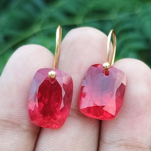 Könnte beinhalten: Ein Paar Ohrhänger mit facettierten, rechteckigen, durchscheinenden roten Steinen. Jeder Stein hängt an einem dünnen, gebogenen goldfarbenen Metallhaken, mit einem kleinen goldfarbenen Perlenakzent an der Stelle, wo der Stein am Haken befestigt ist.