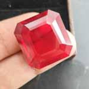 May include: A vibrant, transparent red gemstone with a square-cut shape and faceted edges. The stone exhibits a deep crimson hue and reflects light across its polished surfaces, showcasing a clear, geometric structure with sharp, defined corners.