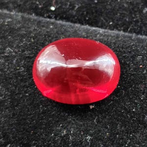 Puede incluir: Una piedra preciosa roja translúcida de forma ovalada con un corte cabujón pulido. La piedra exhibe un tono carmesí profundo y vibrante y una superficie brillante y reflectante que capta la luz, creando un reflejo blanco sobre su parte superior.