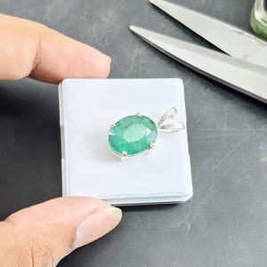 Könnte beinhalten: Ein ovaler, grüner Smaragd-Anhänger in einer silberfarbenen Metallfassung mit Krappen. Der Anhänger hat eine facettierte Oberfläche und eine polierte silberne Öse an der Oberseite und liegt auf einer kleinen, quadratischen weißen Karte.