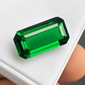 May include: A vibrant emerald green gemstone cut in a rectangular emerald shape with faceted edges. The stone displays high clarity and deep green saturation, reflecting light across its polished surfaces while resting on a white display base.