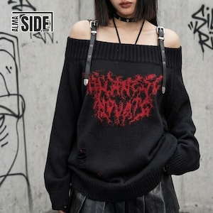 以下が含まれることがあります： 肩出しデザインの黒いニットセーター。ダメージ加工が施された質感で、長袖、リブ編みのネックラインが特徴です。肩にはシルバーのバックルが付いた黒いフェイクレザーのストラップが2本取り付けられており、中央には赤いグラフィックプリントが施されています。