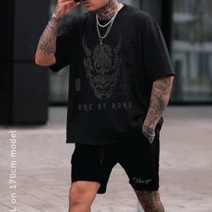 Peut inclure: Un t-shirt noir à manches courtes orné d'un motif blanc représentant un masque Hannya japonais, le texte "ONE OF NONE" et des caractères japonais. Il est associé à un short noir portant un petit logo blanc sur la jambe gauche.