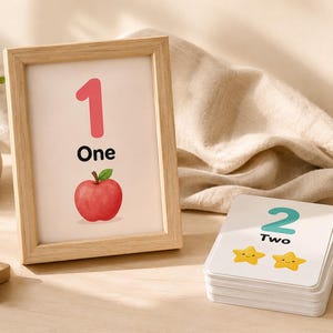 May include: A framed print with the number 1, the word "One," and a red apple illustration. A stack of cards shows the number 2, the word "Two," and two yellow star illustrations. Educational materials for children.