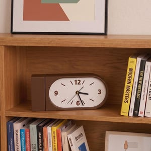 Può includere: Un orologio analogico ovale marrone con quadrante bianco, lancette nere e numeri marroni nelle posizioni 3, 6, 9 e 12. L'orologio è posizionato su uno scaffale in legno accanto a libri intitolati Modern Masters, The New Abstract, Art & Tech e Color Theory.