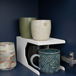 Puede incluir: Una colección de tazas y tazas de cerámica en varios colores y texturas. Las tazas se exhiben en una estantería blanca. Una taza es de color verde azulado con un diseño de huella de pata y hojas.