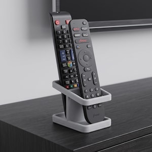 Puede incluir: Un soporte de plástico gris claro para mandos a distancia diseñado para almacenar dos dispositivos en posición vertical. El soporte tiene una base rectangular y un marco de dos niveles, con un mando negro con teclado numérico y otro con el texto free.