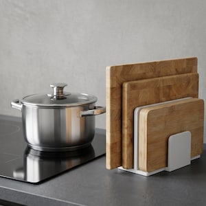 Puede incluir: Una olla de acero inoxidable con tapa de cristal está sobre una placa de cocina negra, junto a un juego de tablas de cortar de madera en un soporte blanco. Las tablas son de diferentes tamaños y de madera clara.