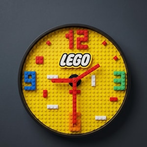 Può includere: Un orologio da parete rotondo fatto di mattoncini LEGO gialli. Il quadrante presenta numeri rossi al 12 e un 9 blu. Il logo LEGO è centrato. Le lancette rosse e nere indicano l'ora. L'orologio è in una cornice nera.