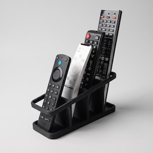 May include: A black plastic remote control organiser with four vertical slots. The organiser holds a black Fire TV remote, a white remote, a black remote with a red power button, and a tall black remote with numerous buttons and a silver top edge.