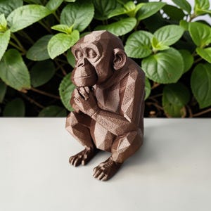 Könnte beinhalten: Eine braune, geometrische Schimpansen-Skulptur in sitzender Position, mit gefalteten Händen vor dem Gesicht. Die Skulptur hat ein facettiertes Design und steht vor einem Hintergrund aus grünem Laub. Der Schimpanse ist ein dekoratives Objekt.