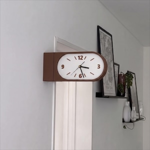 Pode incluir: Um relógio de parede de face dupla com moldura castanha e mostrador oval branco. O relógio apresenta numerais castanhos nas posições das 3, 6, 9 e 12 horas, com ponteiros castanhos para horas, minutos e segundos. Está montado numa parede.