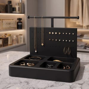 Op de afbeelding: Een matzwarte sieradenhouder met een T-vormige stang voor kettingen, een geperforeerd achterpaneel voor oorknopjes en een basis met vier uitsparingen voor ringen en armbanden. De houder bevat diverse gouden en zilveren sieraden.
