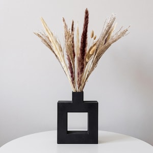 Può includere: Un vaso quadrato nero opaco con un'apertura centrale, contenente una composizione di erbe e piume essiccate nei toni del crema, beige e bordeaux scuro. Gli steli botanici essiccati si aprono a ventaglio dal collo stretto del vaso geometrico.