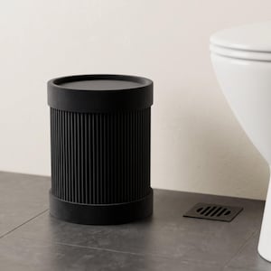 Pode incluir: Um cesto de lixo cilíndrico preto com uma textura verticalmente estriada na sua superfície exterior. O recipiente tem um rebordo superior circular liso e plano e uma base sólida a condizer, apresentando um acabamento mate uniforme.