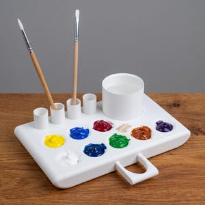Può includere: Una tavolozza per pittura in plastica bianca con dieci scomparti circolari riempiti con vari colori di vernice acrilica, un grande bicchiere cilindrico per l'acqua e quattro portapennelli. Due pennelli sono inseriti nei supporti.