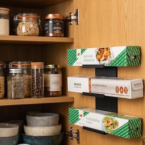 Pode incluir: Um suporte de parede de metal preto contendo três caixas retangulares de suprimentos de cozinha. A caixa superior está rotulada como Aluminium-Allzweckfolie, a do meio como Migros Kitchen & Co. Backpapier Rolle e a inferior como Frischhaltefolie.