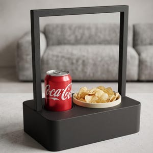 Op de afbeelding: Een matzwarte rechthoekige snackhouder met een hoog handvat, voorzien van een ronde uitsparing voor een rood Coca-Cola blikje en een beige kom gevuld met lichtgekleurde aardappelchips, ontworpen voor handig serveren op tafel.