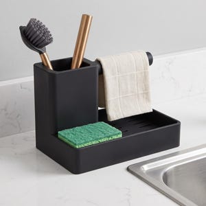 Op de afbeelding: Een matzwarte gootsteenorganizer met een hoog vierkant vak voor een grijze afwasborstel en een metalen hulpmiddel, een horizontale stang met een beige geblokte doek en een platte basis met een rechthoekige groene spons.