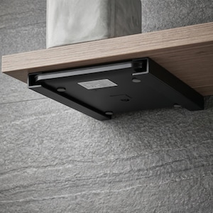 Op de afbeelding: Een zwarte, rechthoekige weegschaal is onder een houten plank gemonteerd. De weegschaal heeft een label en kleine, ronde voetjes. Een betonnen object staat op de plank erboven. De plank is bevestigd aan een grijze stenen muur.