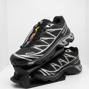 Pode incluir: Um par de ténis de trail running Salomon XT-6 pretos e prateados com membrana Gore-Tex, sistema de atacadores rápidos com detalhes em amarelo e vermelho e uma sola intermédia cinzenta. Os sapatos estão empilhados num suporte preto.