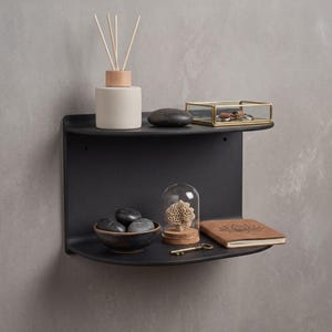 Peut inclure: Une étagère murale noir mat à deux niveaux incurvés. Le niveau supérieur contient un diffuseur à roseaux blanc, une pierre sombre lisse et une boîte en verre rectangulaire à cadre doré. Le niveau inférieur contient un bol sombre de pierres, une cloche en verre avec un objet corallien, une clé en métal et un carnet beige avec un motif de fleur de lotus.