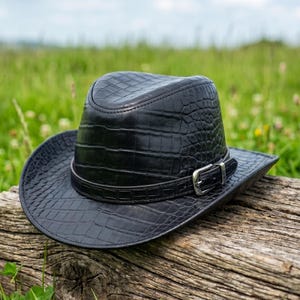 Puede incluir: Un sombrero de vaquero negro con un patrón texturizado similar al de un cocodrilo. El sombrero tiene una hebilla plateada en una banda de cuero. Descansa sobre un tronco de madera desgastado, con un fondo de hierba verde y un cielo azul.
