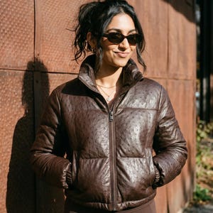 Puede incluir: Una chaqueta acolchada de color marrón oscuro con un patrón texturizado de piel de avestruz, que presenta un cuello alto, una cremallera frontal completa y mangas largas con puños elásticos. La chaqueta tiene una construcción acolchada.