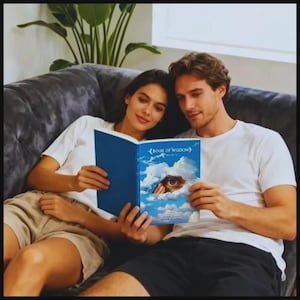 May include: A couple is seated on a gray couch, reading a book together. The book has a blue cover with an eye peeking through clouds. The book's title is "Book of Wisdom". The couple is wearing white t-shirts.