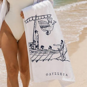 May include: A white beach towel featuring a black line art illustration of a classical Greek ship with figures on board and a large bird flying above. The Greek text OΔYΣΣEIA is printed in black near the bottom edge of the rectangular fabric.