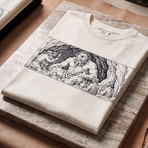 May include: An off-white t-shirt featuring a black ink-style illustration of Polyphemus in a cave. The graphic depicts a cyclops with figures holding spears and vessels. The text "POLYPHEMUS IN THE CAVE" is printed in small capital letters at the bottom.