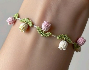May include: A delicate crochet bracelet featuring a floral design. The bracelet showcases small, three-dimensional flowers in shades of pink, white, and yellow, interspersed with green leaves. The bracelet is worn on a wrist.
