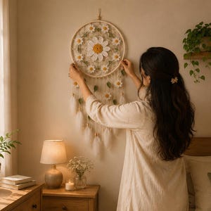 Puede incluir: Un atrapasue&ntilde;os con tem&aacute;tica de margaritas con un aro color crema y flores de ganchillo blancas y amarillas. El atrapasue&ntilde;os tiene plumas y cuentas colgantes. La imagen muestra el atrapasue&ntilde;os colgado en una pared.