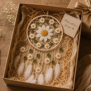 Puede incluir: Un atrapasue&ntilde;os hecho a mano con un dise&ntilde;o de margarita, presentado en una caja de regalo. El atrapasue&ntilde;os presenta una gran margarita central y margaritas m&aacute;s peque&ntilde;as alrededor del aro, con plumas blancas y el texto "Made with love for you".