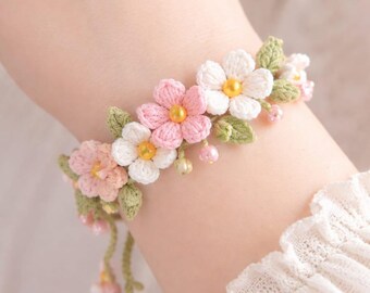 May include: A handmade crochet bracelet featuring pink and white flowers with yellow centers and green leaves. The floral design is accented with small pink beads. The bracelet is a delicate piece of jewelry.