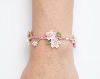 May include: A pink crochet bracelet adorned with delicate pink and white flowers and green leaves. The bracelet is secured with a silver clasp and is worn on a wrist. The floral design adds a touch of nature-inspired elegance.