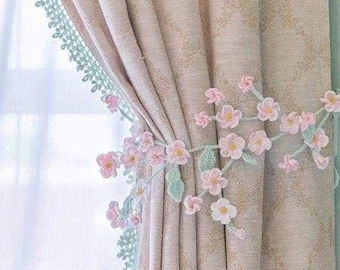 May include: A beige curtain with a subtle diamond pattern, featuring a light green crocheted lace trim along the edge and a decorative tie-back adorned with numerous small, light pink crocheted flowers and pale green crocheted leaves.