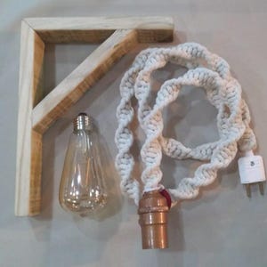 May include: A wooden corner shelf with a light bulb and a macrame cord. The light bulb is clear with a copper-colored base. The macrame cord is white and has a plug at the end. The wooden shelf is light brown.