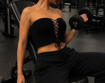 May include: A black, knitted tube top with a lace-up front is worn with black sweatpants. A 7.5 kg dumbbell is being lifted in a gym setting. A black water bottle with the word "PROTEIN" is visible.