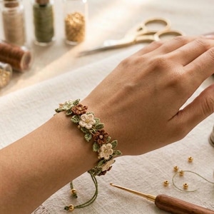 Può includere: Braccialetto floreale fatto a mano con fiori all'uncinetto color crema, marrone e verde. Il braccialetto è su un polso, con un cordino verde e perline dorate. Forniture per l'artigianato, tra cui forbici e filo, sullo sfondo.