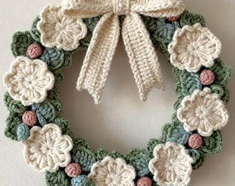 May include: A circular crochet wreath featuring a base of dark green leaves, accented with small light blue and pink spherical berries, and decorated with several cream-colored crocheted flowers. A large cream-colored crochet bow hangs from the top.