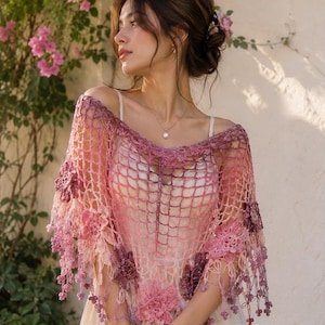 Puede incluir: Un chal de crochet hecho a mano, en tonos rosas y morados. El chal presenta adornos florales y flecos con pequeñas cuentas. La prenda se lleva sobre un vestido blanco, creando una estética delicada y femenina. Perfecto para una ocasión especial.